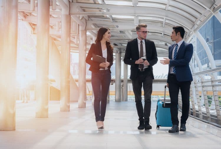 Geschäftsreisende auf dem Weg zum Meeting – lohnrelevante Abrechnung von Dienstreisen Drei Personen in Businesskleidung gehen mit einem Rollkoffer durch ein modernes Gebäude – typische Szene für lohnbuchhalterisch relevante Dienstreiseabrechnungen