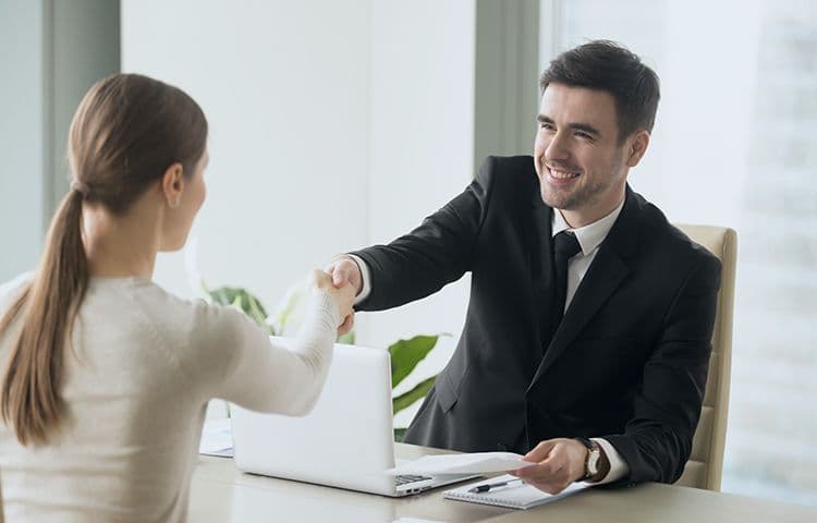 Handschlag zwischen Bewerberin und Personalverantwortlichem im Büro Ein Mann im Anzug schüttelt einer Frau die Hand in einem modernen Büro. Beide lächeln. Auf dem Tisch liegen ein Laptop und Unterlagen. Die Szene deutet auf ein Bewerbungsgespräch oder Vertragsabschluss hin.