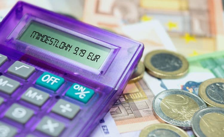 Taschenrechner mit Anzeige „Mindestlohn 9,19 EUR“ auf Euro-Geldscheinen und Münzen Lila Taschenrechner mit der Anzeige „Mindestlohn 9,19 EUR“, daneben Euro-Geldscheine und Münzen – Symbol für Lohnberechnung und Gehaltsabrechnung