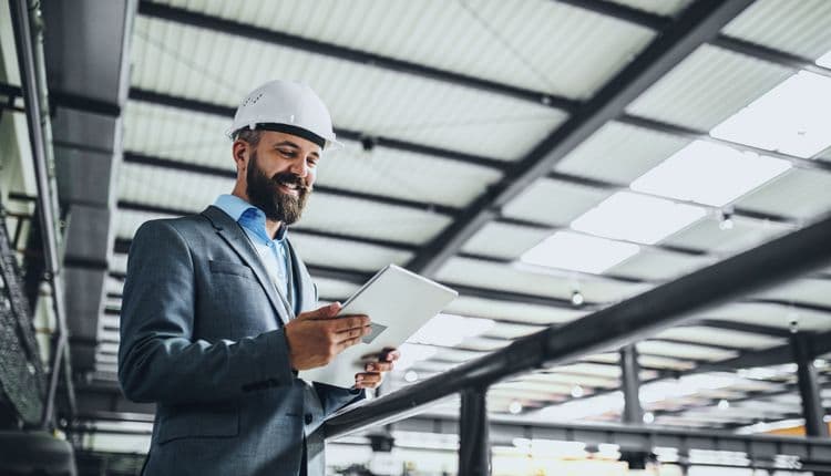 Gewerblicher Mitarbeiter mit Tablet – digitale Lohnbuchhaltung im Industriebereich Ein Mann mit Schutzhelm und Anzug steht in einer Industriehalle und arbeitet auf einem Tablet – moderne Lohnabrechnung und Zeiterfassung im gewerblichen Bereich