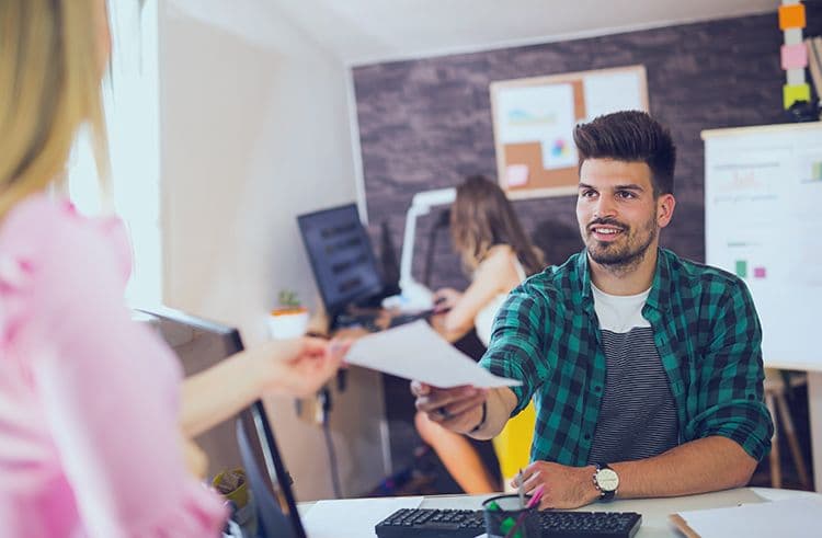 Dokumente für die Lohnabrechnung: Übergabe im modernen Arbeitsumfeld Junger Mitarbeiter nimmt im Büro ein Dokument von einer Kollegin entgegen – Symbol für Lohnabrechnung oder HR-Prozesse