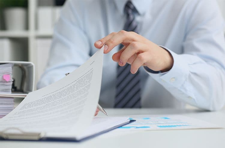 Mann im Hemd blättert durch ein Dokument auf einem Schreibtisch Ein Mann in einem hellblauen Hemd prüft ein Dokument auf einem Clipboard. Auf dem Tisch liegen weitere Unterlagen mit Diagrammen sowie ein Stift. Daneben befindet sich ein geöffneter Aktenordner mit Papierstapeln.