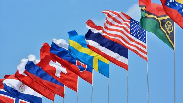 Länderflaggen – Symbol für internationale Lohnabrechnung und Mitarbeiterentsendung Mehrere Länderflaggen, darunter USA, Schweden, Schweiz und Norwegen, wehen nebeneinander im Wind vor blauem Himmel