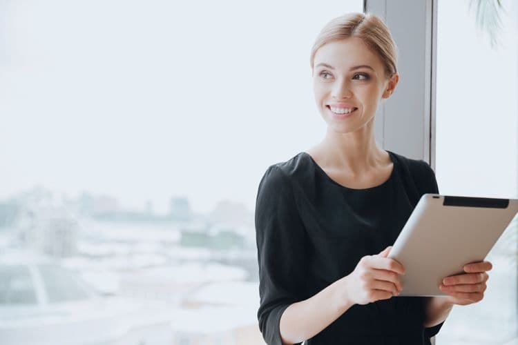 Lächelnde Frau mit Tablet vor Fenster im Büro – Lohnbuchhaltung Junge Frau im schwarzen Kleid steht lächelnd mit einem Tablet in der Hand vor einem großen Fenster mit Blick auf eine Stadtlandschaft – symbolisch für moderne Lohnbuchhaltung oder Personalverwaltung