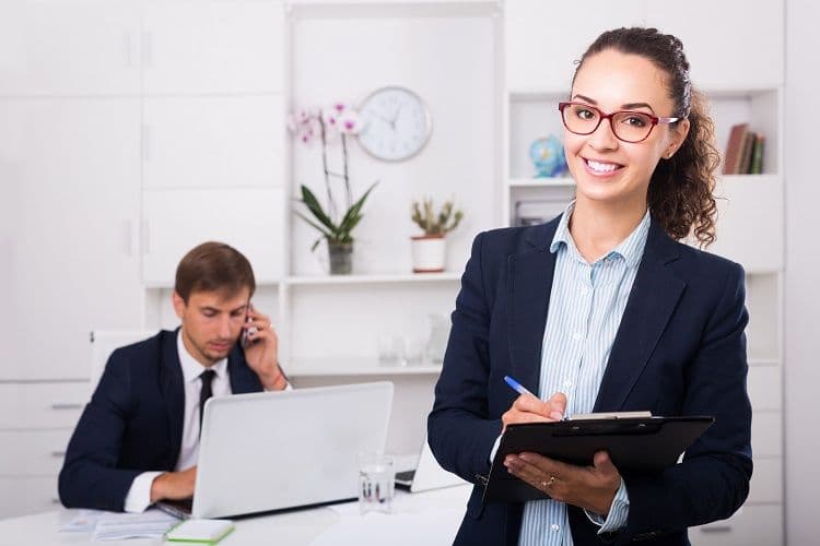 Zwei Büroangestellte bei der Arbeit – Teamarbeit in der Lohnbuchhaltung Eine junge Frau mit Brille steht im Vordergrund mit einem Clipboard und einem Stift, während ein Kollege im Hintergrund telefoniert und am Laptop arbeitet. Das Bild zeigt ein professionelles Umfeld, das typisch für die Lohnbuchhaltung ist.